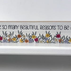May include: A white wooden sign with a floral border and the text "There are so many beautiful reasons to be happy".