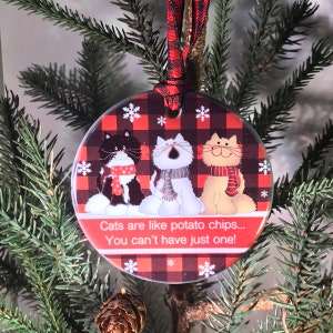 May include: Round Christmas ornament featuring three cartoon cats wearing scarves, set against a red and black plaid background with snowflakes. The ornament has a red and black ribbon for hanging, and the text reads "Cats are like potato chips... You can't have just one!"