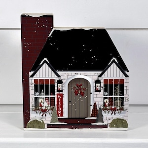 May include: A white and red wooden Christmas house decoration with a black roof, a red brick chimney, and a welcome sign. The house has two windows with snow-covered window boxes and a wreath on the door.