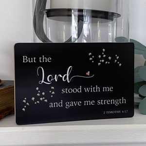 May include: Black wooden sign with white text that reads "But the Lord stood with me and gave me strength 2 Timothy 4:17". The sign has a small heart and scattered green and pink dots.