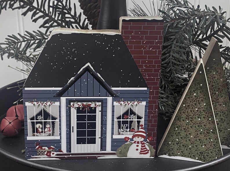 Op de afbeelding: Een set van drie houten kerstversieringen met een blauw huis met een rode bakstenen schoorsteen, een sneeuwpop met een rode muts en sjaal, en een groene kerstboom. Het huis heeft een witte deur en ramen met rode bekleding. De sneeuwpop houdt een rood hart vast en de boom is bedekt met witte sneeuw.