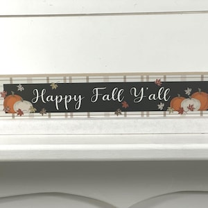 May include: A rectangular wooden block sign featuring a beige and white plaid border. The center is a black horizontal panel with the white cursive text Happy Fall Y'all, decorated with illustrations of orange and white pumpkins and autumn leaves.