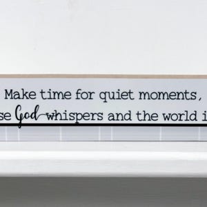 May include: White wooden sign with a black and white quote that reads "Make time for quiet moments, because God whispers and the world is loud." The sign is decorated with a green branch with two small yellow birds.