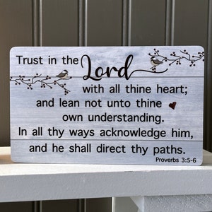 May include: A wooden sign with a whitewashed background and a gray woodgrain pattern. The sign features a bible verse from Proverbs 3:5-6, which reads "Trust in the Lord with all thine heart; and lean not unto thine own understanding. In all thy ways acknowledge him, and he shall direct thy paths." The sign is decorated with two small birds and a heart.