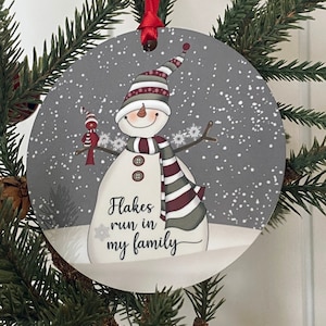 May include: Round Christmas ornament with a snowman and a cardinal. The snowman is wearing a red and white striped hat and a green and red striped scarf. The background is gray with white snowflakes. The text "Flakes run in my family" is written below the snowman.