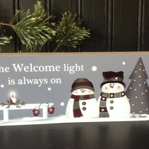 May include: A rectangular wooden sign with a gray background features the text "The Welcome light is always on." Two snowmen wearing hats and scarves stand next to a gray Christmas tree. A small bench with a candle and mittens is also depicted.