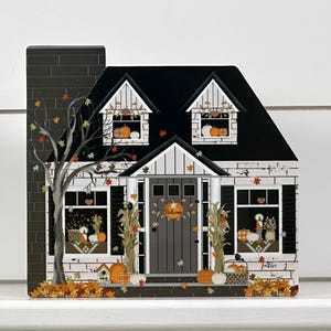 May include: A decorative wooden house with a black roof, chimney, and door. The house is decorated with pumpkins, fall leaves, and the word "Welcome" on the door. A tree with fall leaves is on the left side of the house.
