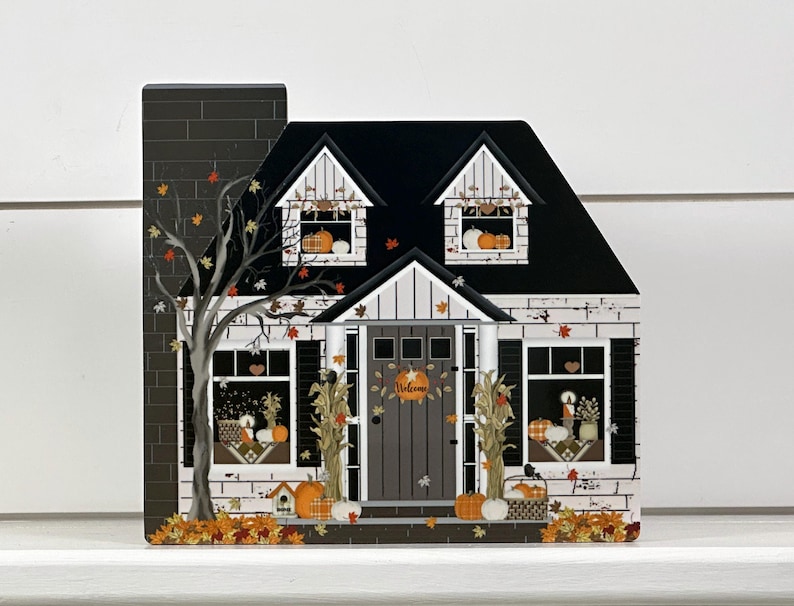 May include: A decorative wooden house with a black roof, chimney, and door. The house is decorated with pumpkins, fall leaves, and the word "Welcome" on the door. A tree with fall leaves is on the left side of the house.
