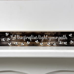 May include: Wooden sign with a rustic woodgrain background and white text that reads "Let the fireflies light your path". The sign is decorated with small white fireflies.