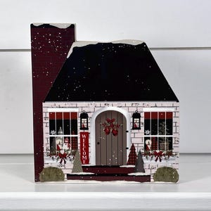 May include: A white and red wooden Christmas house decoration with a black roof, a red brick chimney, and a welcome sign on the front door.