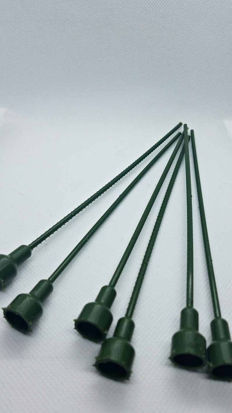 May include: A collection of six dark green floral stem picks. Each pick has a textured stem and a flared, cup-shaped head. The stems are approximately 6 inches long, and the heads have a jagged edge.