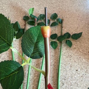 May include: Artificial rose stems with green leaves and a red and green stem. The rose buds are yellow and orange. The stems are green and the leaves are a dark green color. The rose stems are for floral arrangements.