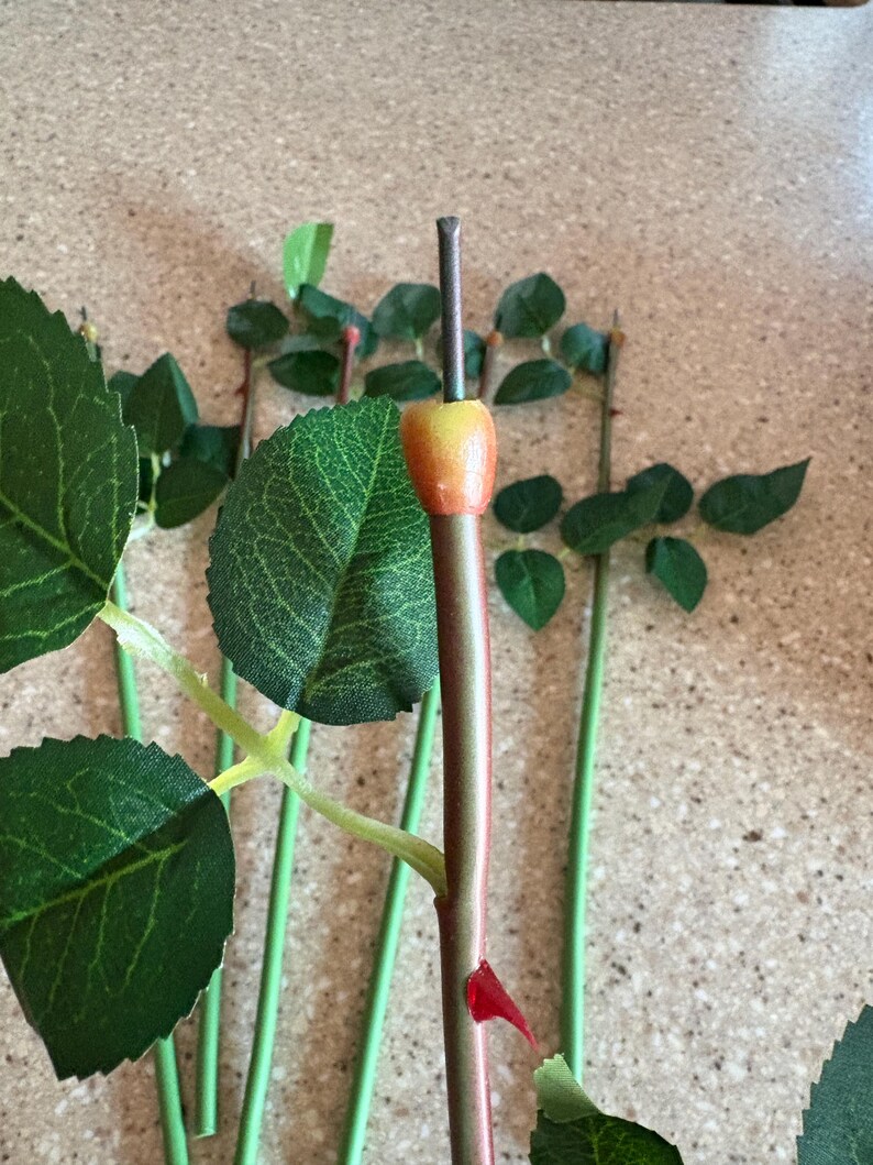 May include: Artificial rose stems with green leaves and a red and green stem. The rose buds are yellow and orange. The stems are green and the leaves are a dark green color. The rose stems are for floral arrangements.