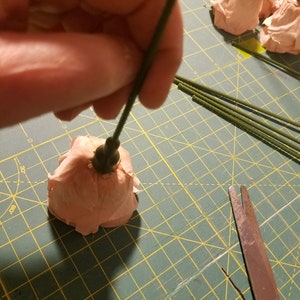 May include: A person is shown assembling a paper flower. The flower is a light peach color, and the stem is green. Other paper flowers and green stems are on a green cutting mat. Scissors are in the foreground.