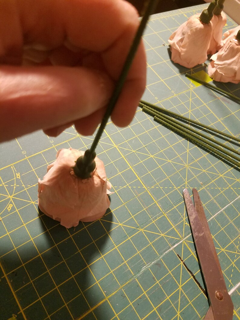 May include: A person is shown assembling a paper flower. The flower is a light peach color, and the stem is green. Other paper flowers and green stems are on a green cutting mat. Scissors are in the foreground.