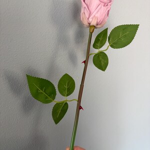 May include: A single artificial rose with a light pink bloom and green leaves. The stem is brown and green with small red thorns. The rose is held in a hand against a gray background.