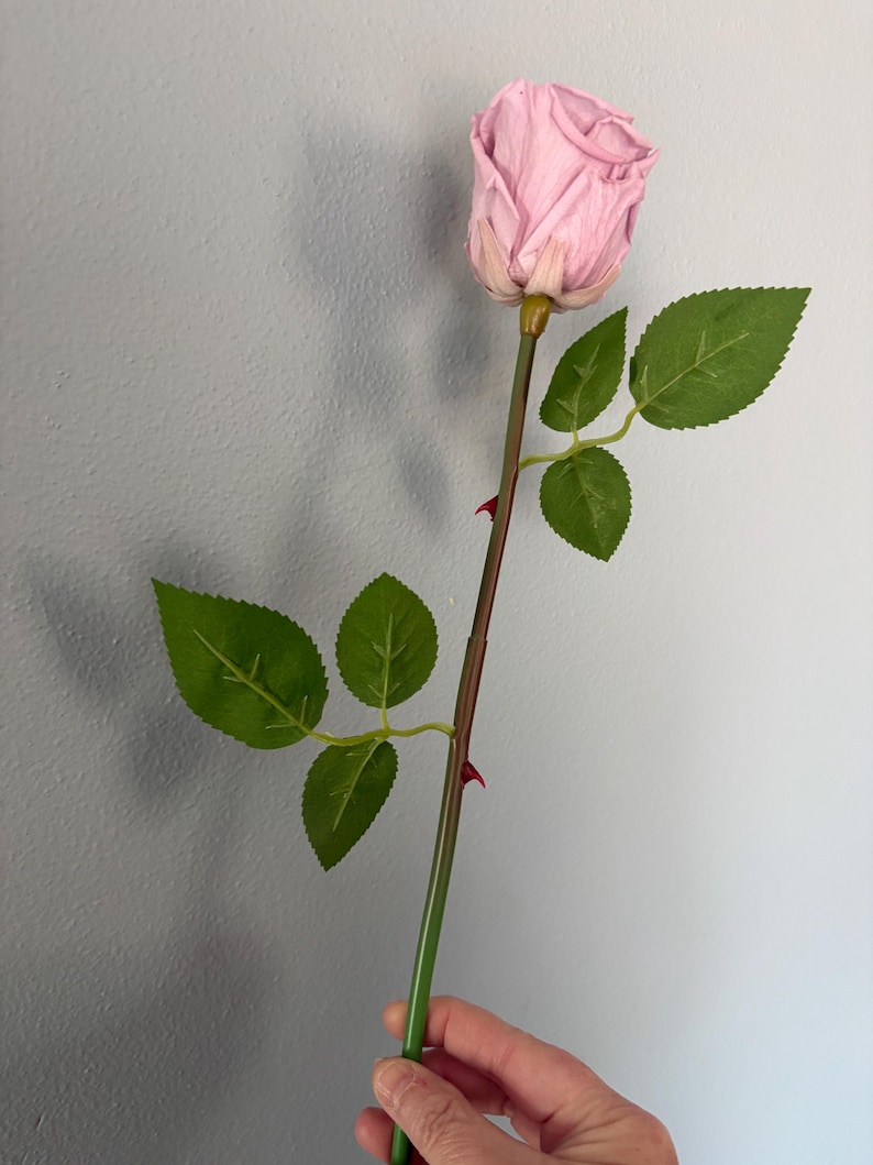 May include: A single artificial rose with a light pink bloom and green leaves. The stem is brown and green with small red thorns. The rose is held in a hand against a gray background.