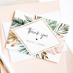 May include: A white thank you card with a gold diamond frame and tropical leaf design. The text "Thank you" is written in black script font. The names "Olivia + Jason" are written below the text.