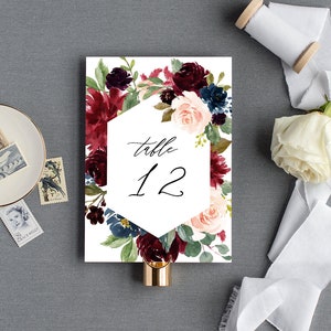 May include: A white wedding table number card with a burgundy, blush, and navy floral design. The card features a hexagonal frame with the text "table 12" in black calligraphy.