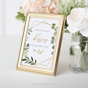 May include: Gold framed sign with a white background and green foliage. The sign says "Guess how many kisses for the soon-to-be Mrs."