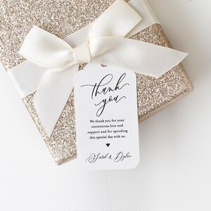 May include: A white thank you tag with black calligraphy that reads "Thank you". The tag has a small heart and the text "Hotel & Dylan" in black calligraphy. The tag is tied to a gold glitter gift box with a white satin ribbon.