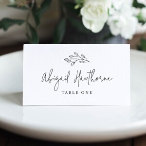 May include: White place card with black calligraphy that reads "Abigail Hawthorne" and "Table One". A small black line drawing of a sprig of leaves is above the name.