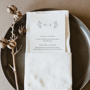 May include: A white folded napkin with a printed menu card on top. The menu card has a simple floral design and the text "G+L" at the top. The menu lists a "First Course" of Cornish Crab Bisque with poached scallops and parsley toasts and a "Second Course" of salad of greens with roasted butternut squash, toasted hazelnuts, goat cheese, and a sherry vinaigrette.