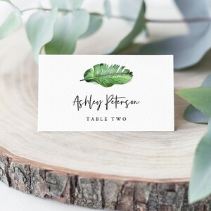 May include: White place card with a green tropical leaf illustration. The text "Ashley Peterson" and "Table Two" is printed in black.
