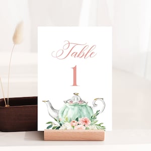Könnte beinhalten: Eine weiße Karte mit dem Text "Tisch 1" in rosa Schrift. Unter dem Text befindet sich eine Aquarell-Illustration einer grünen Teekanne mit rosa und weißen Blumen. Die Karte steht auf einem Holzständer.