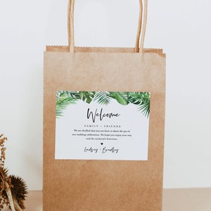 May include: A brown paper gift bag with a welcome sign. The sign has a tropical leaf border and says "Welcome Family & Friends. We are thrilled that you are here to share the joy of our wedding celebration. We hope you enjoy your stay and the weekend's festivities. Lindsey & Bradley"