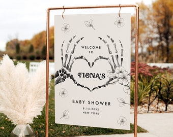 May include: A copper-colored metal frame holds a sign with the words "Welcome to Fiona's Baby Shower." The sign features a heart made of skeleton hands, floral accents, and the date and location. A vase of pampas grass is in the foreground.