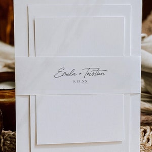 May include: White wedding invitation with a simple design. The invitation has three layers and a band with the text "Emile + Tristan 9.15.XX" in black script.
