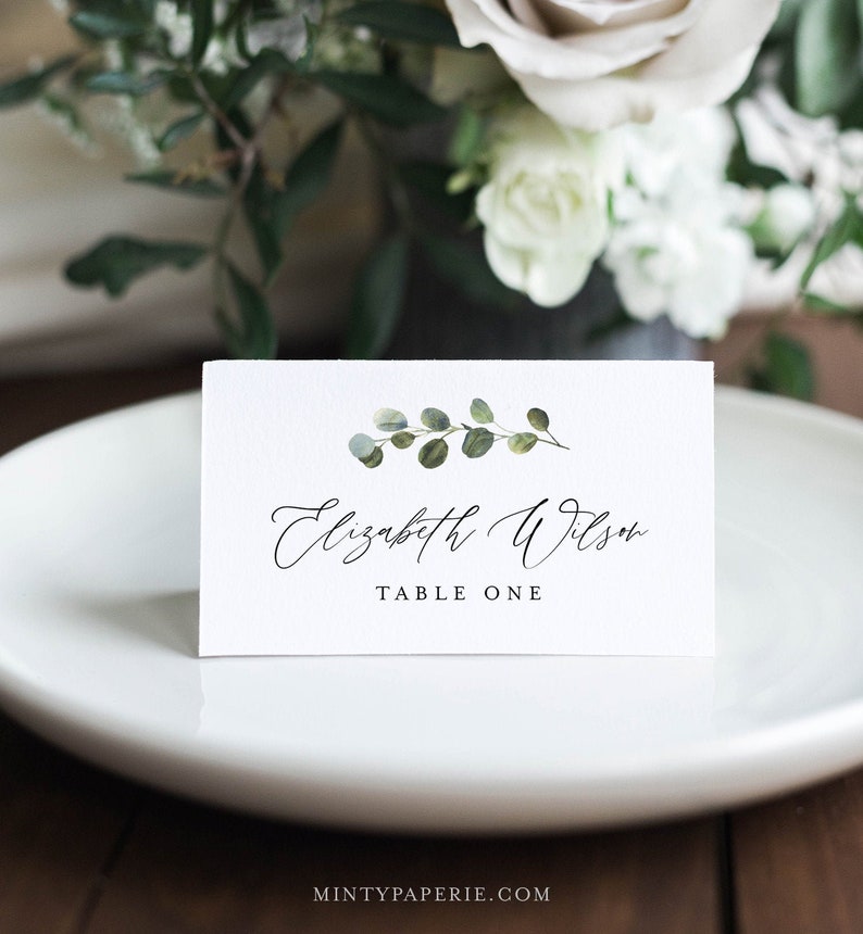 May include: White place card with a green eucalyptus sprig illustration and the text "Elizabeth Wilson" and "Table One" in black.