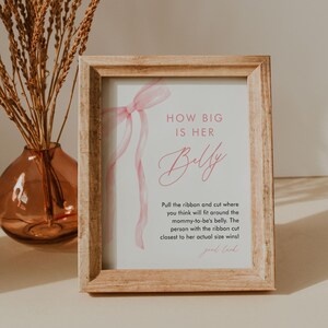 May include: A framed sign with the text "HOW BIG IS HER Belly" in pink script. The sign includes instructions for a baby shower game, with a pink ribbon design. The frame is made of light-colored wood.