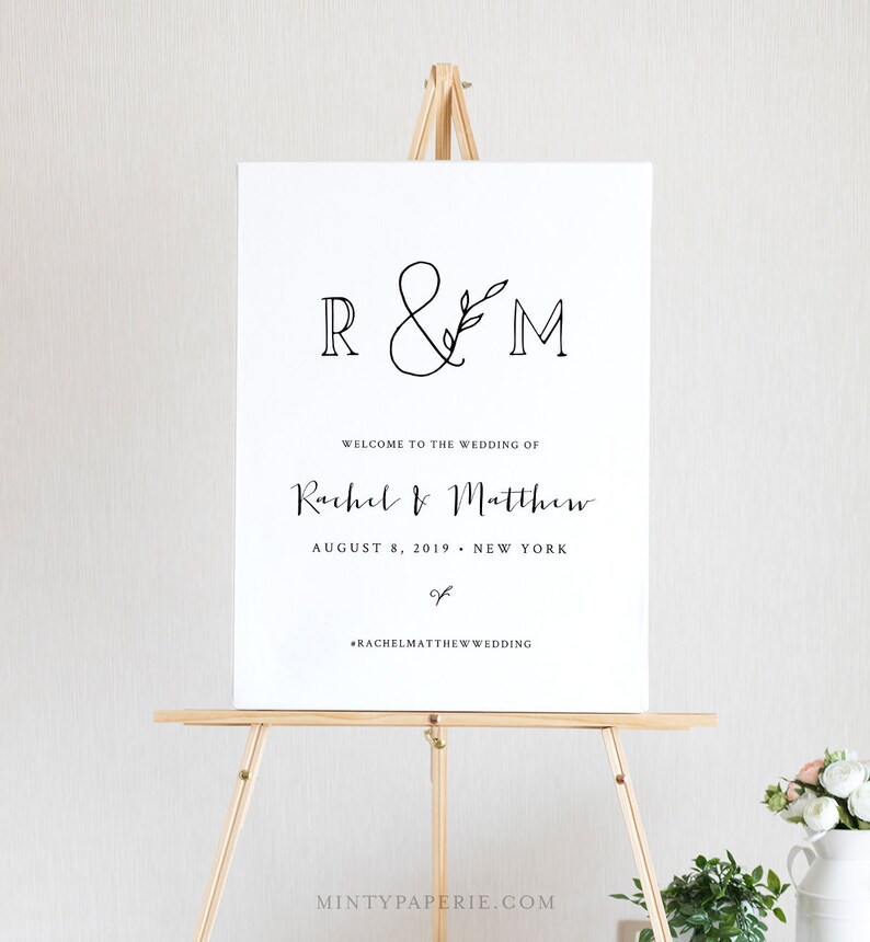 May include: A white wedding welcome sign with black text and a simple floral design. The sign reads "R&M" with a leafy branch, "Welcome to the wedding of Rachel & Matthew", "August 8, 2019 - New York", and "#RachelMatthewWedding".