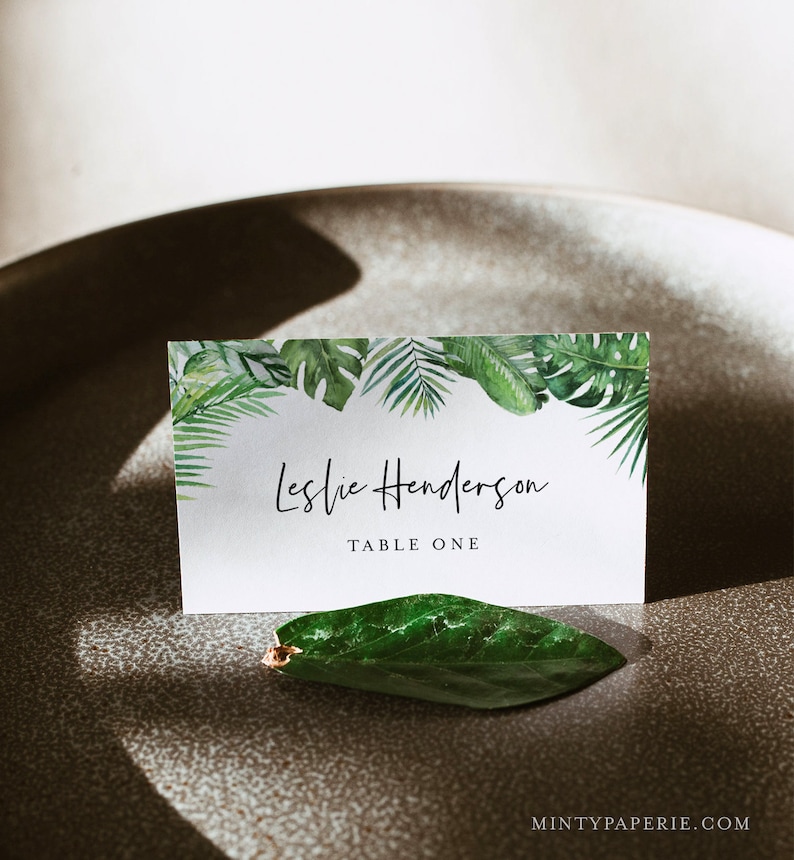 May include: A white place card with a tropical leaf border. The text "Leslie Henderson" is written in black cursive script. The text "Table One" is written in black sans-serif font.