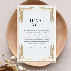 May include: A white thank you card with gold art deco style borders and text. The card reads "THANK YOU" at the top, followed by a message of gratitude and the names "HARPER + ADRIAN" at the bottom. The card is on a wooden plate.