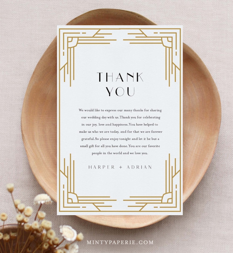 May include: A white thank you card with gold art deco style borders and text. The card reads "THANK YOU" at the top, followed by a message of gratitude and the names "HARPER + ADRIAN" at the bottom. The card is on a wooden plate.