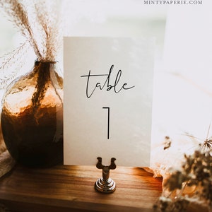 Puede incluir: Tarjeta de n&uacute;mero de mesa blanca con caligraf&iacute;a negra que dice "mesa 1" sobre una superficie de madera con un soporte plateado.
