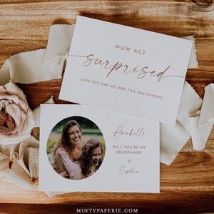Può includere: Due biglietti bianchi con testo color oro rosa e una foto di due donne. Il biglietto in alto recita "NOW ACT surprised (LIKE YOU HAD NO IDEA THIS WAS COMING)". Il biglietto in basso chiede "Anabelle WILL YOU BE MY BRIDESMAID?" con il nome Sophia. Il sito web mintypaperie.com è in basso.