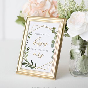May include: A gold framed sign with a white background and green foliage border. The sign says "Guess how many kisses for the soon-to-be Mrs."