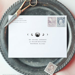 May include: A white envelope with a moon phase design, addressed to Mr. and Mrs. Anderson, rests on a silver platter. Vintage stamps and gold scissors are also on the platter. The envelope includes the address: 501 Willow Boulevard, Westwood, NJ 07690.