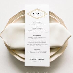 May include: A rectangular menu card with a gold geometric design at the top, featuring the word "MENU". The menu lists a multi-course meal, including Cornish Crab Bisque, Roasted Leg of English Lamb, and a Glazed Lemon Tart. The card is on a beige plate with a folded napkin.