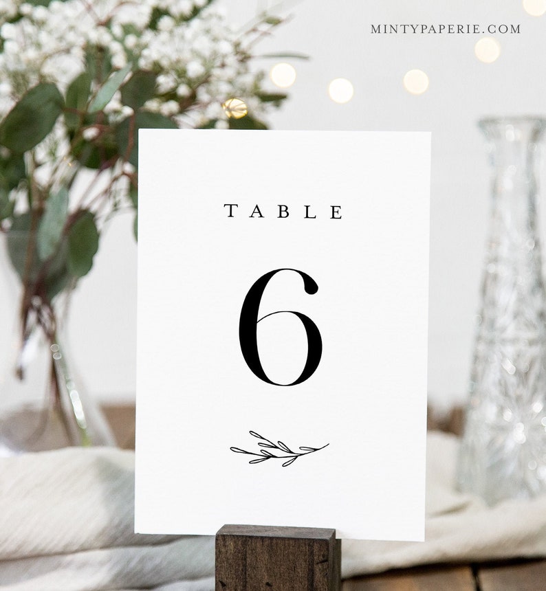 May include: White table number card with black text that reads "TABLE 6" with a small black floral design at the bottom.