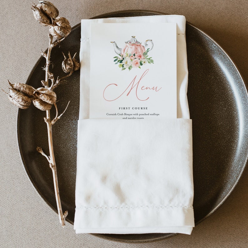 May include: A white napkin with a menu card tucked inside. The menu card features a watercolor illustration of a teapot with flowers and the word "Menu" in cursive. The first course listed is "Cornish Crab Bisque with poached scallops and toasted croutons."
