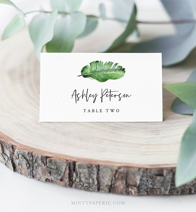May include: White place card with a green tropical leaf illustration. The text "Ashley Peterson" and "Table Two" is printed in black.