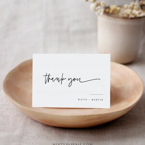 May include: White thank you card with black calligraphy that says "thank you" and the names "Katie - Martin" printed below.