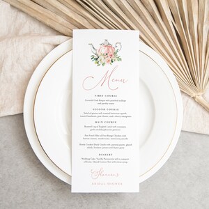 May include: A white menu card with a pink teapot illustration and floral accents. The menu lists a four-course meal for a bridal shower, including Cornish Crab Bisque, Salad of Greens, Roasted Leg of English Lamb, and Wedding Cake.