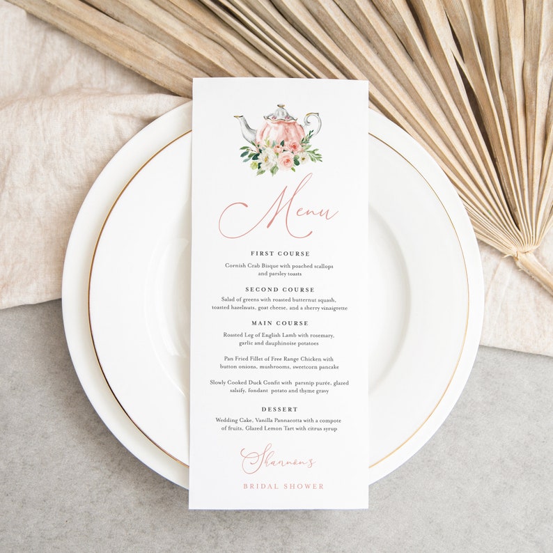 May include: A white menu card with a pink teapot illustration and floral accents. The menu lists a four-course meal for a bridal shower, including Cornish Crab Bisque, Salad of Greens, Roasted Leg of English Lamb, and Wedding Cake.