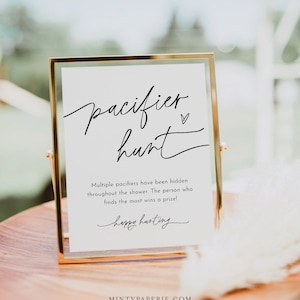May include: A white sign in a gold frame with black text that reads "pacifier hunt". The sign says "Multiple pacifiers have been hidden throughout the shower. The person who finds the most wins a prize!" and "happy hunting".